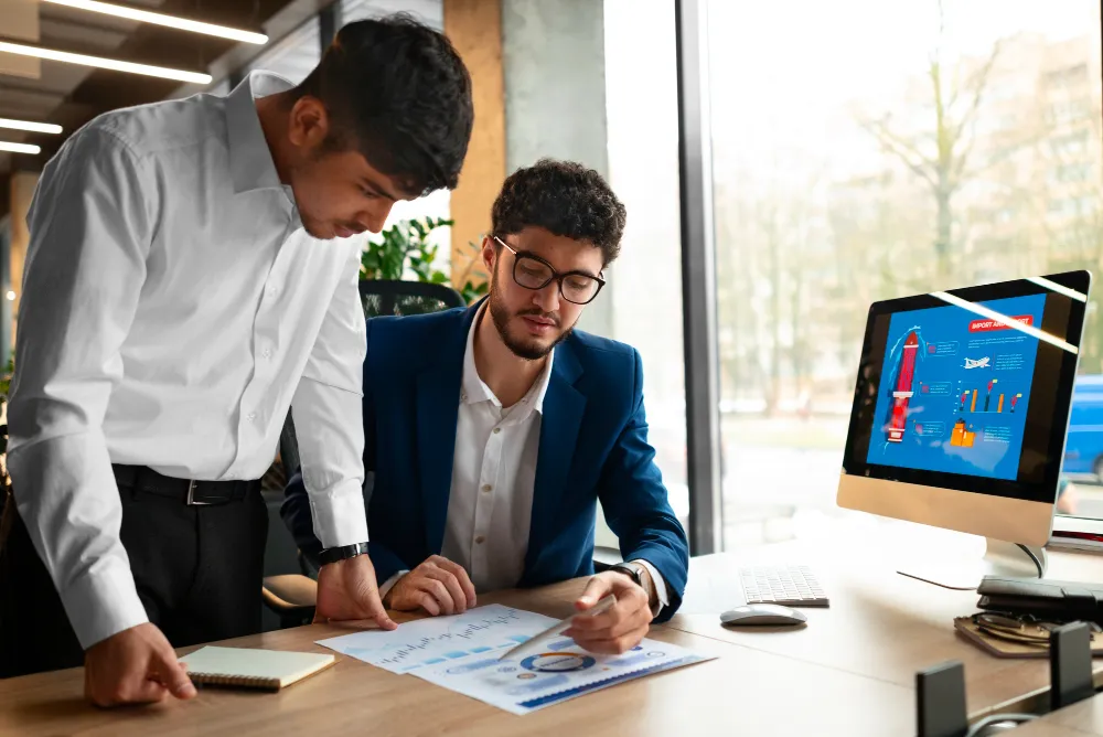 Two professionals analyzing business reports and data charts in a modern office, reviewing performance insights displayed on a computer screen.